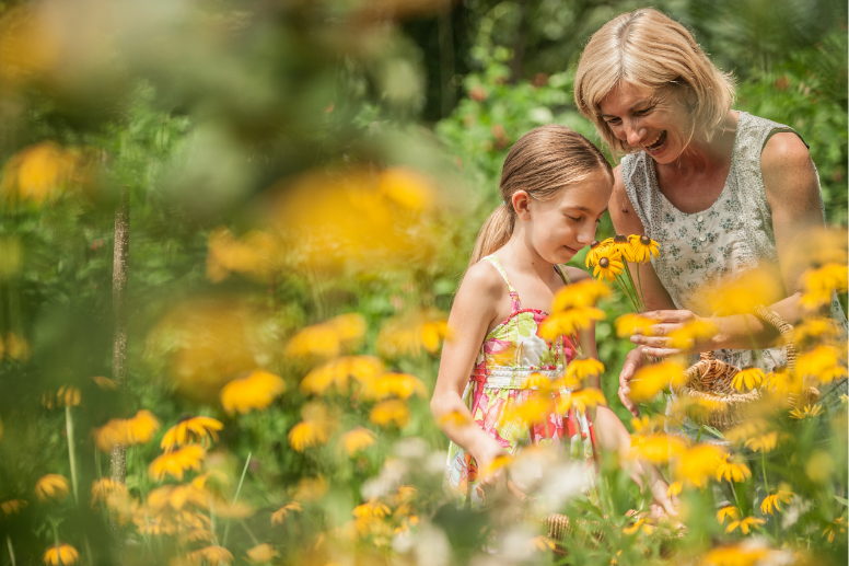 Mutter und Tochter stehen auf einer Blumenwiese, mit gelben Blumen und riechen an einer Blüte.
